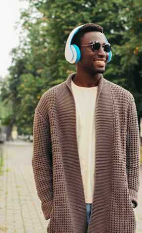 A man in a brown coat and sunglasses is wearing headphones and standing on a sidewalk.