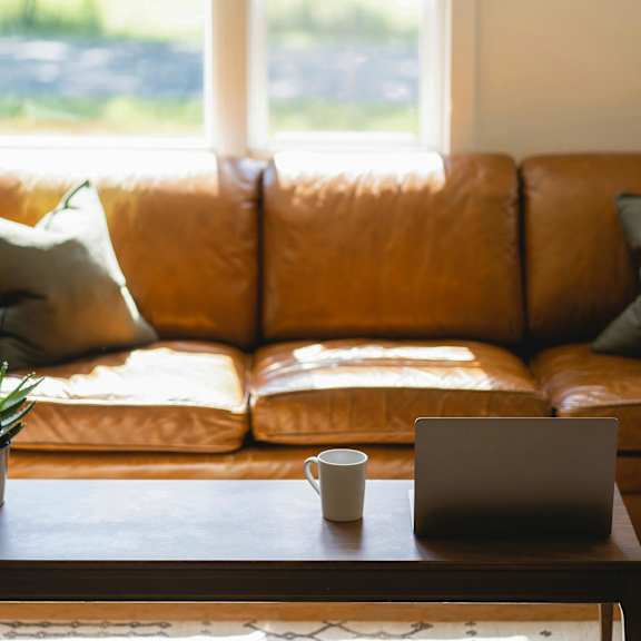 A brown leather couch with sage green pillows and a coffee table with a mug and laptop on it.