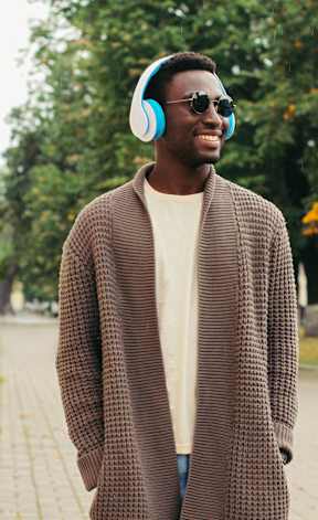 A man in a brown coat and sunglasses is wearing headphones and standing on a sidewalk.