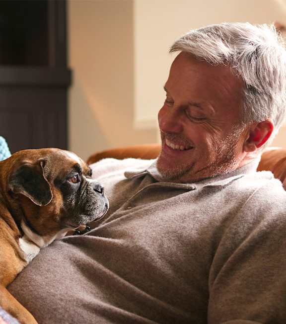 A man is sitting on a couch with a dog and holding a mug.