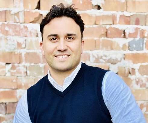 A man in a blue vest and light blue shirt is sitting in front of a brick wall.