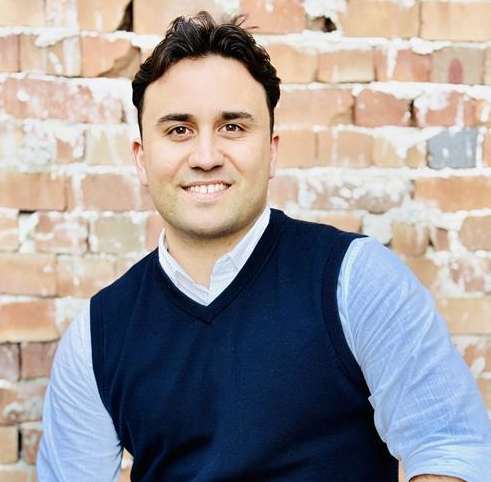A man in a blue vest and light blue shirt is sitting in front of a brick wall.
