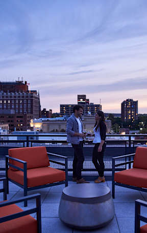 Two people standing on a rooftop terrace with orange chairs and a city skyline in the background.