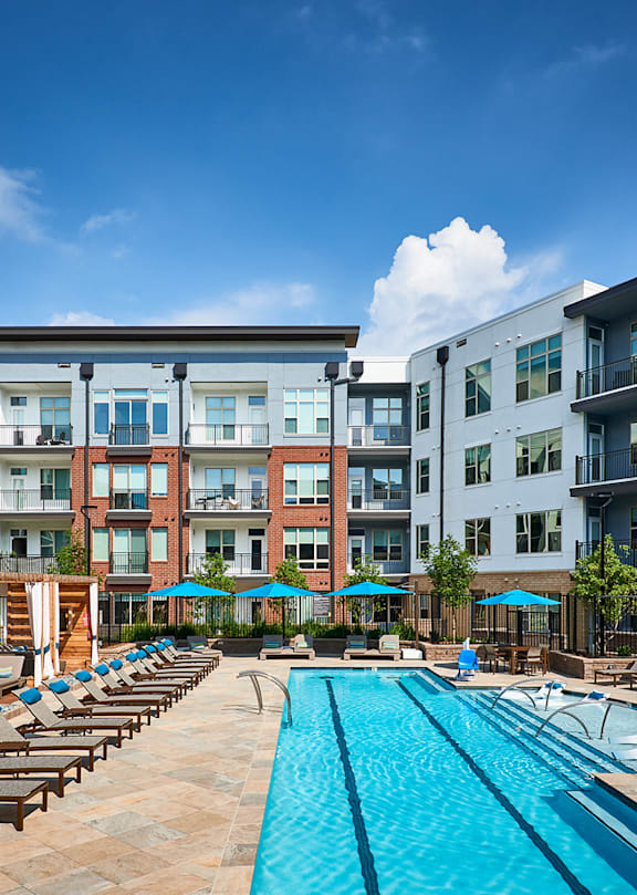 A large swimming pool surrounded by lounge chairs and umbrellas in front of apartment buildings.