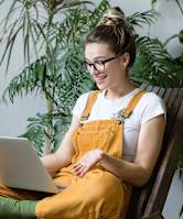 A woman in a yellow jumpsuit is sitting on a chair with a laptop in front of her.