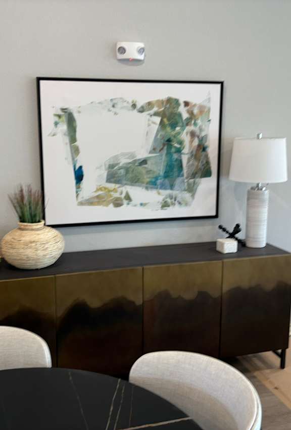 A black and white painting hangs above a black cabinet with a white vase and a white lamp on top.