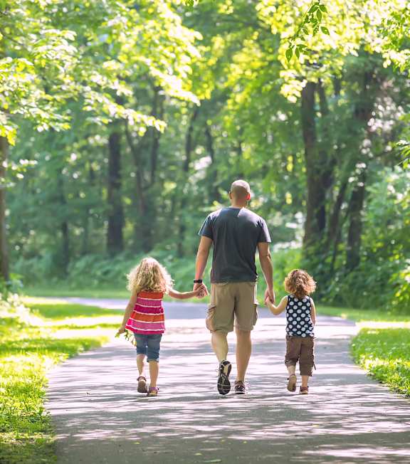 A man and two children are walking down a path in a green forest at Poplar Glen Apartments, Columbia, Maryland.