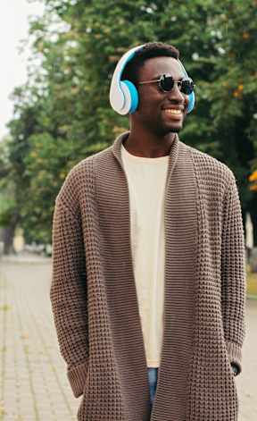 A man in a brown coat and sunglasses is wearing headphones and standing on a sidewalk.