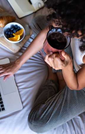 A woman sitting on a bed drinking coffee and eating breakfast on the computer at Eastpark Apartment Homes, Salem, OR