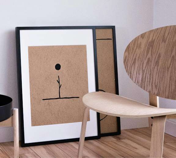 A framed artwork with a brown background and a black border is on a wall next to a round wooden table with a candle and a vase with reeds.