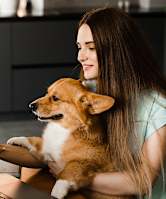 A woman is sitting at a desk with a laptop and a phone, holding a corgi.