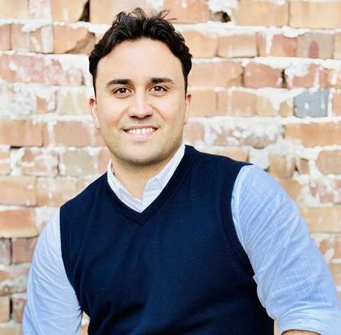 A man in a blue vest and light blue shirt is sitting in front of a brick wall.