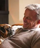 A man is sitting on a couch with a dog and holding a mug.