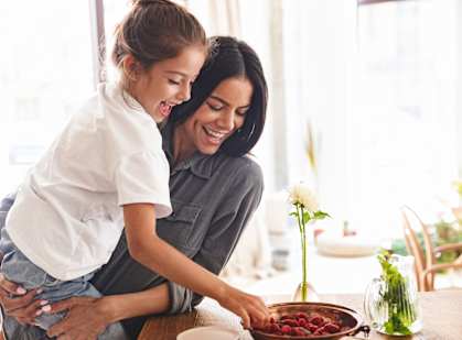 A woman and a young girl are sitting at a table with a bowl of strawberries in front of them.