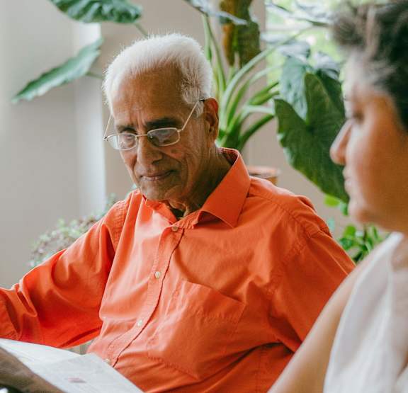 An elderly man in an orange shirt is reading a newspaper while a woman in a white shirt looks on.