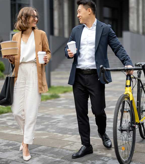A man in a suit walks with a bicycle while holding a cup and a woman in a coat walks behind him.