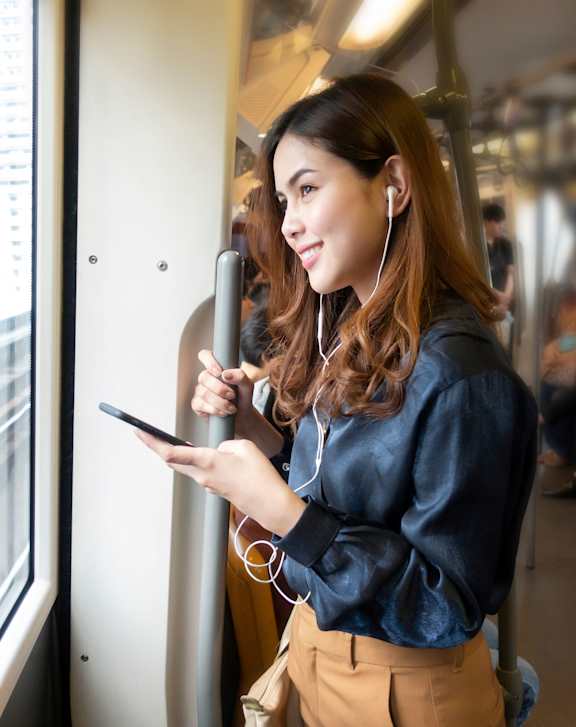 A woman in a blue blouse and beige pants is looking at her phone while standing on a train.