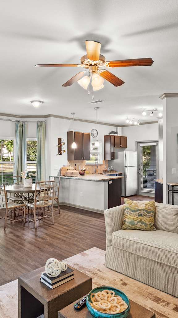 An open apartment layout with a living room ceiling fan, a dining area with bay windows, and a fully-equipped gourmet kitchen.