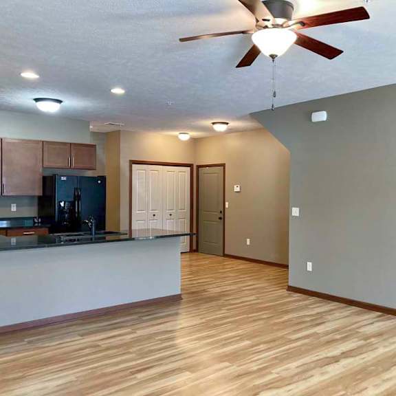 A living room with hardwood-style flooring and a ceiling fan, open to a modern kitchen with brown cabinets, dark granite counters, and black appliances.