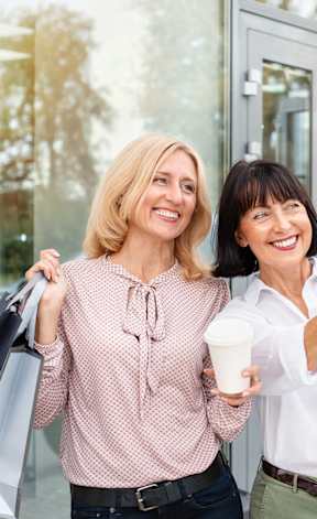 Two women with shopping bags and a cup of coffee are pointing at something outside.