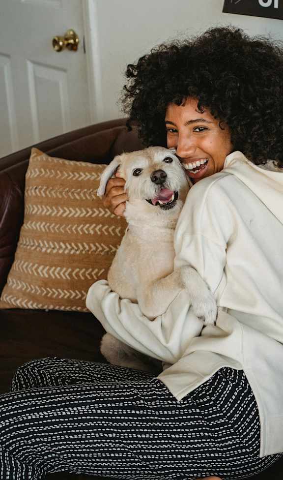 A woman with curly hair is sitting on a brown leather couch holding a white dog.