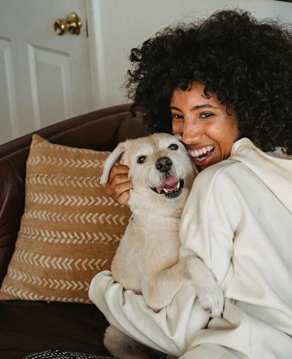 A woman with curly hair is sitting on a brown leather couch holding a white dog.