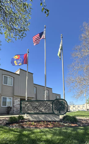 Park 25 Apartments signage near the front entrance of the leasing office, with a tall building, flags, and trees in the background.