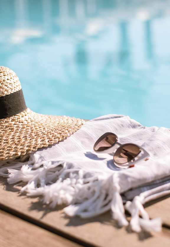 A straw hat, sunglasses, and a towel are placed on a wooden table by a pool.