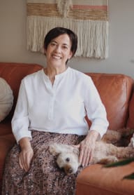 A woman in a white shirt and patterned skirt sits on a brown couch with a small dog. at The Platform, Kent, 98032