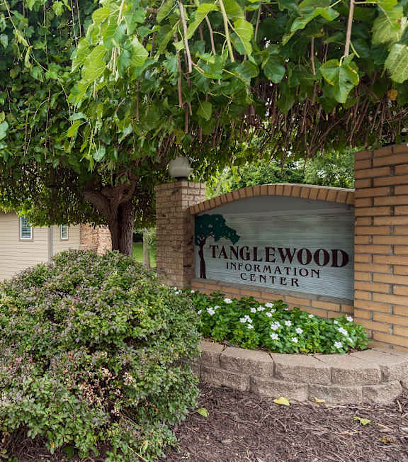 A welcoming Tanglewood sign marks the entrance to an apartment complex, surrounded by lush green bushes and framed by mature trees.