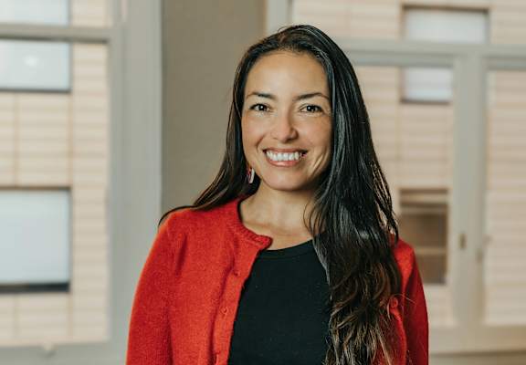 A woman with long dark hair wearing a red cardigan and black top standing in front of a window.