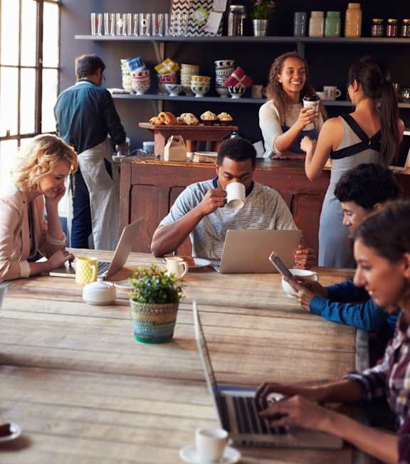 A group of people are sitting around a table in a café, using laptops and drinking coffee.