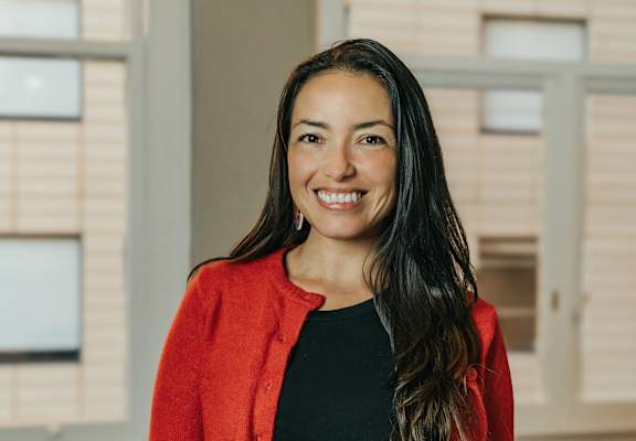 A woman with long dark hair wearing a red cardigan and black top standing in front of a window.