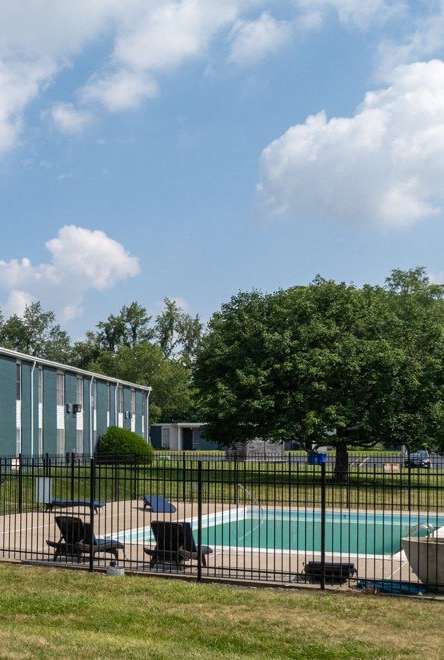 A swimming pool is surrounded by a fence and chairs in front of a building.