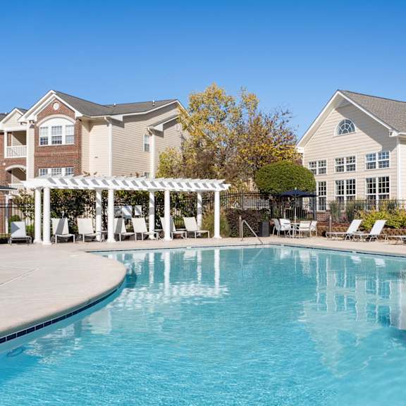 A large swimming pool in front of a building with a white pergola and lounge chairs. at Village on the Lake Apartments, Apartments, Spring Lake, NC