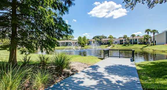 A wooden walkway leads to a pond in a sunny park.