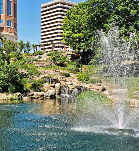 A fountain in the middle of a pond surrounded by trees and buildings.