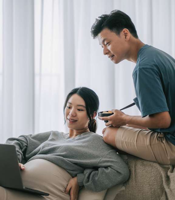 A man is sitting on a couch with a woman, both looking at a laptop screen.