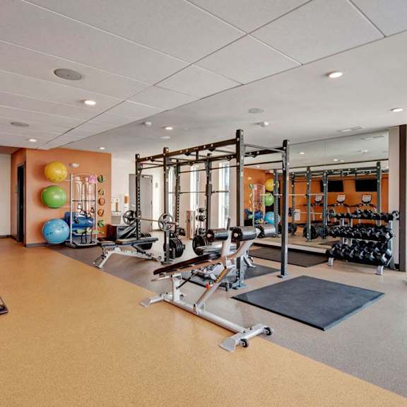 A spacious, bright gym with orange walls, cardio machines, windows, TVs, exercise balls, and free weights at Stitchweld Apartment Homes. 