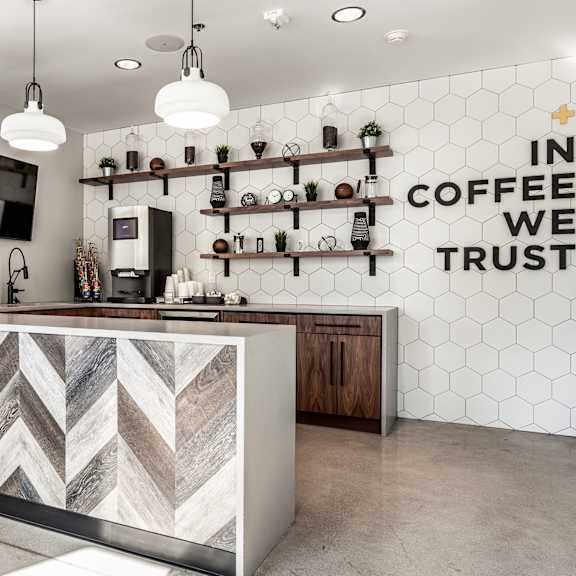 The community kitchen with a coffee machine, toaster, fridge, marble counters, a TV, and a sign that says "In Coffee We Trust" at Vim + Vigor Lofts. 