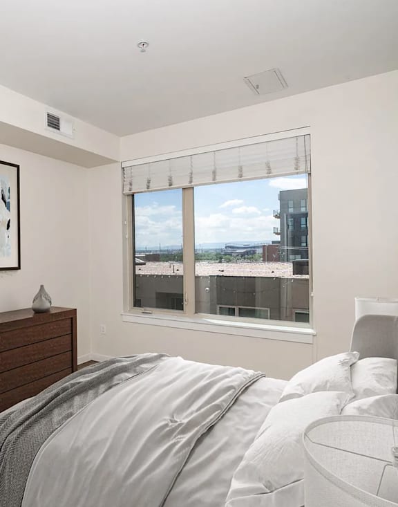 A bedroom with a bed, nightstand, and a window overlooking a cityscape.