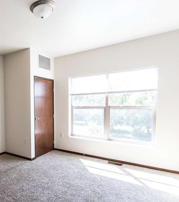 A spacious room with a grey carpet and a window letting in natural light.