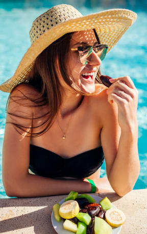 woman wearing sun hat and sunglasses in a pool eating fruits