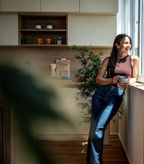 A woman in a brown top and blue jeans stands by a window in a modern kitchen.