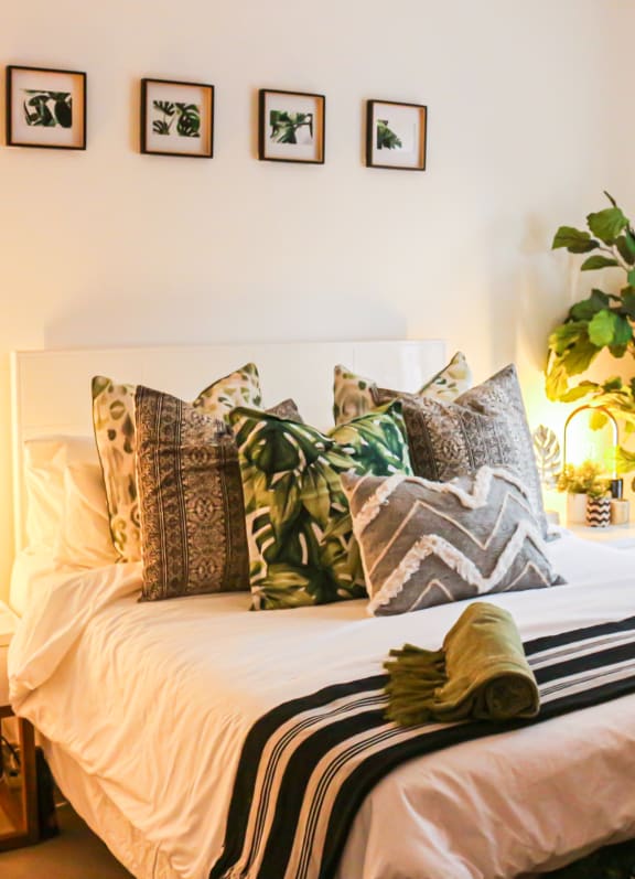 Bedroom with grey and green throw pillows. The bedding is white and black. There is a fig tree next to the bed to the right. Above the bed are 4 pieces of artwork.