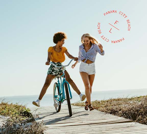Two people riding a bicycle on a wooden path near the beach.