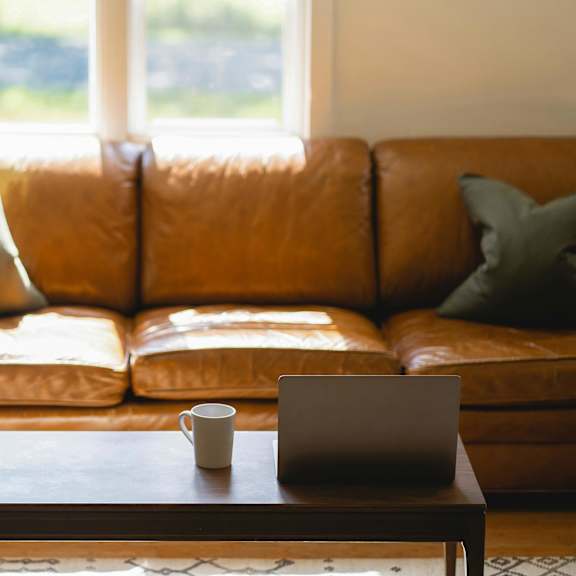 A brown leather couch with a laptop and a cup on a wooden table in front of it.