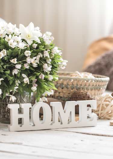 A white wooden table with a white flower pot, a white HOME sign, and a brown knitted pillow.
