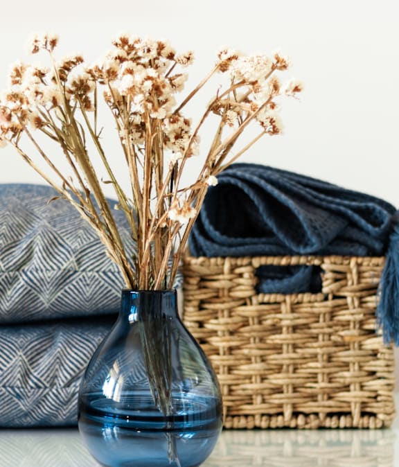 A blue vase with dried flowers sits on a table next to a wicker basket and two patterned pillows.