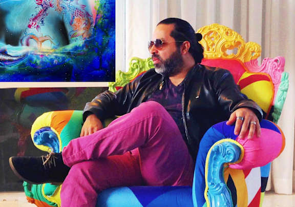 A man in a black leather jacket and pink pants is sitting on a colorful chair in front of a painting.